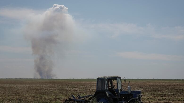 A tractor is seen in a field as smoke rises from a military strike in the background.