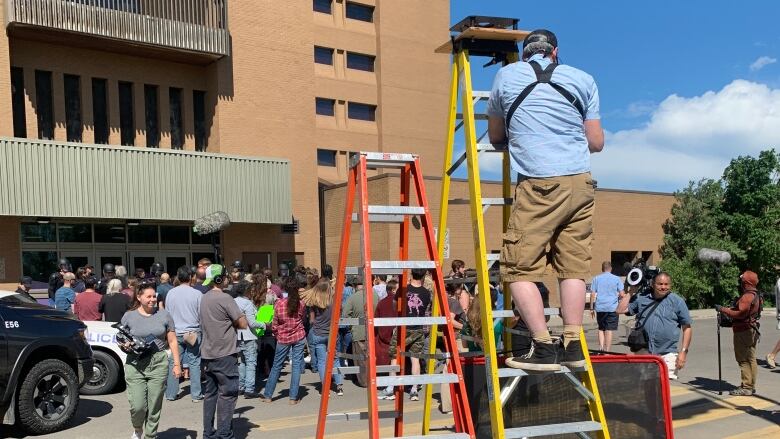 A movie set with a man on a ladder with a camera in front of a crown of people who are doing a scene holding signs. 