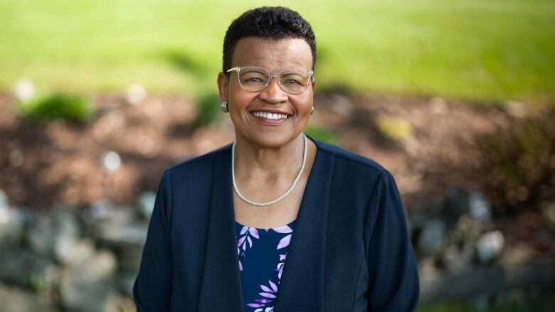 A smiling woman poses for a photograph. She is wearing glasses and a necklace, and a navy blazer with a blouse underneath. 