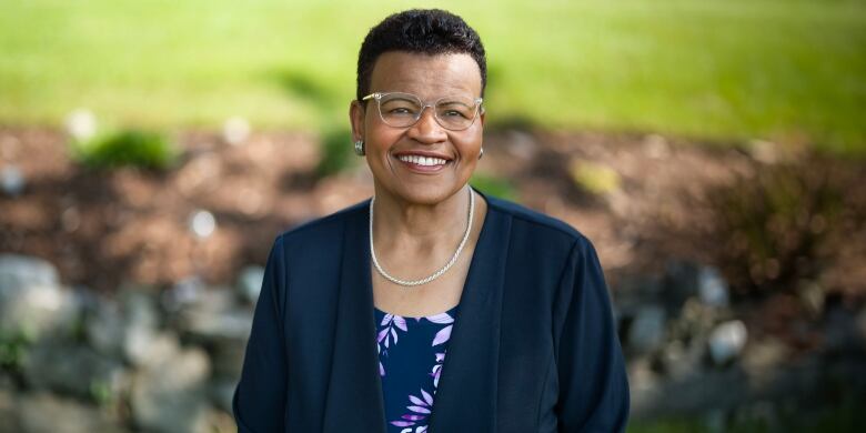 A smiling woman poses for a photograph. She is wearing glasses and a necklace, and a navy blazer with a blouse underneath. 