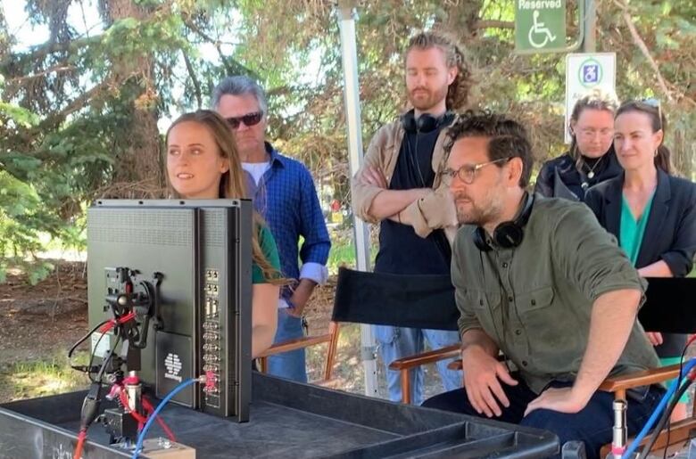 A director sits in a chair behind a screen watching with other people from the crew around him. 