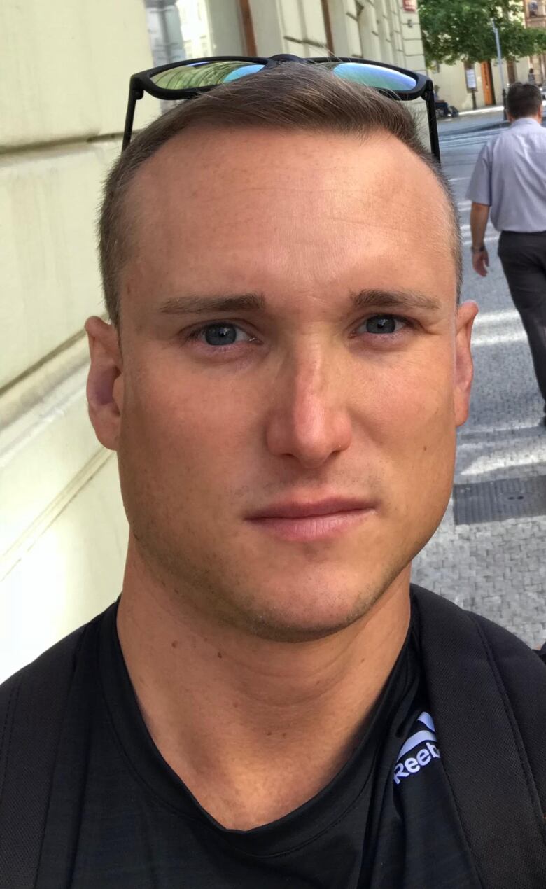 A man in a black shirt with sunglasses on his head stands in the street. 