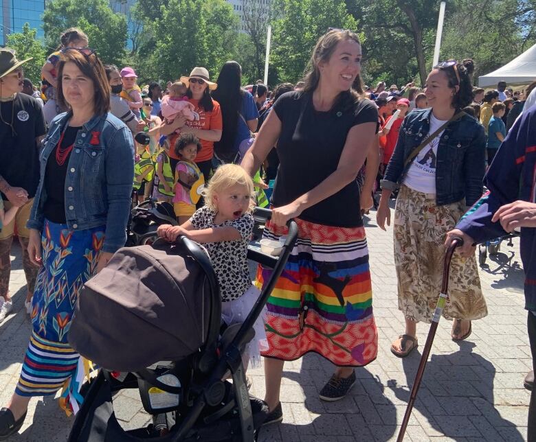 A woman in a blue ribbon skirt and a woman in a rainbow ribbon skirt, pushing a stroller, walk on the street.