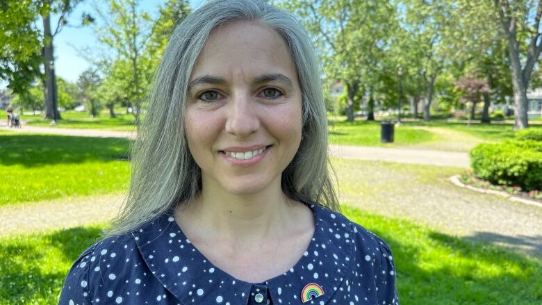 A woman with shoulder-length grey hair in a blue dress with a rainbow Pride pin smiling in a park with bright green trees and grass in the background.