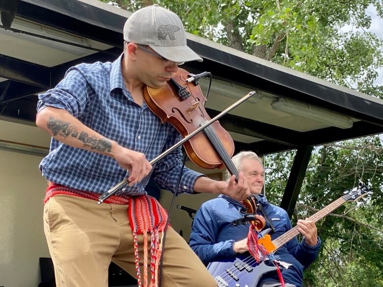 A man wearing a Metis sash plays the fiddle, and behind him another man plays an electric guitar.