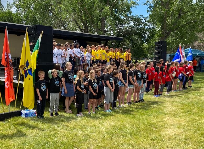 Four groups of students sing in a choir formation, one group wearing black shirts, one group wearing white shirts, one group wearing yellow shirts, and one group wearing red shirts.