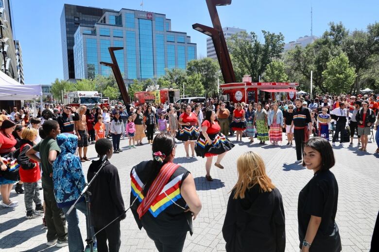 Dancers in Metis clothes, with red shirts and black striped skirts, stand inside of a large circle of people.