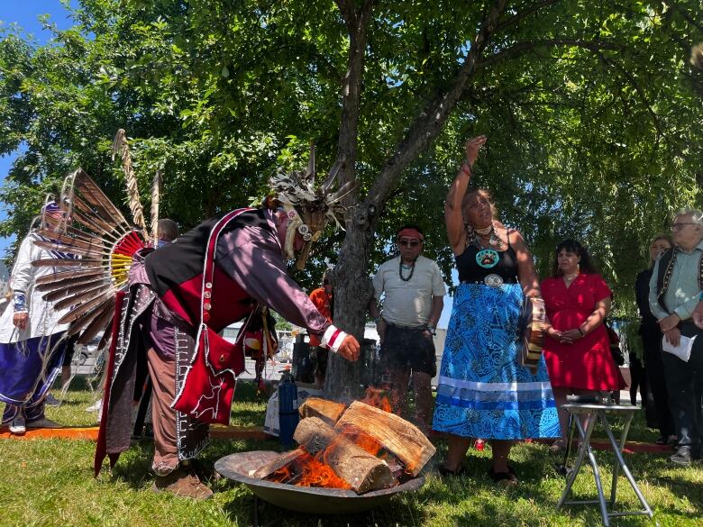 A man wearing traditional Mohawk regalia throws tobacco into a fire with a woman in a blue skirt and Bear Clan necklace next to him. 