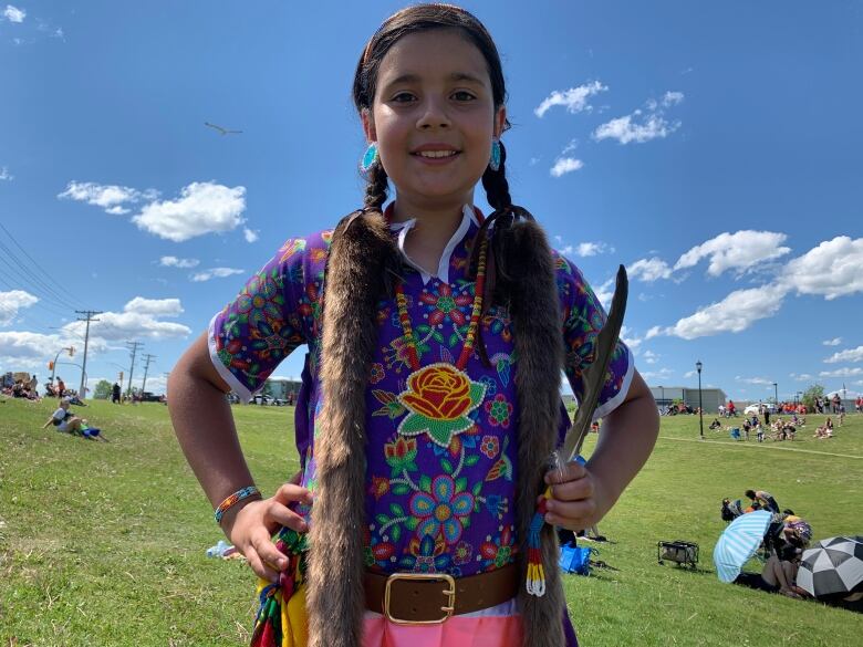 A girl in regalia holds her hands on her hips with an eagle feather in one hand. 