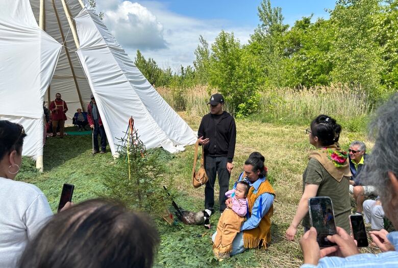 A traditional teepee in a field, with spruce boughs covering the ground outside. A young special needs child dressed in moose hide is helped by his father to walk.