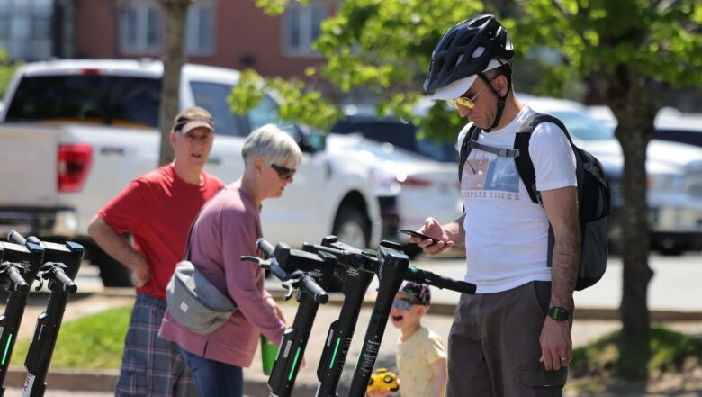 A man in a helmet and sunglasses looks down at his phone while standing in front of a rack of scooters