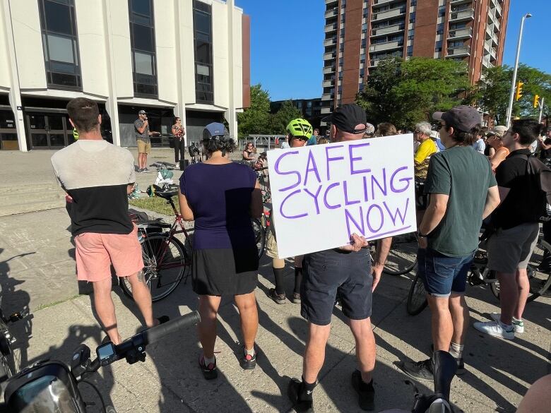 People listen to a speaker at an outdoor plaza in a city. One holds a sign saying SAFE CYCLING NOW.