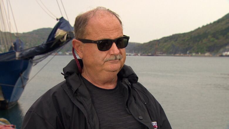 A bald man in a black shirt and black jacket, wearing black RayBan sunglasses. He's standing in front of St. John's harbour.