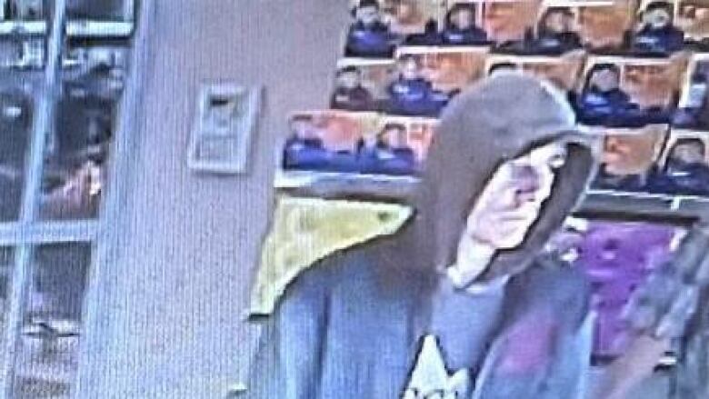 A still image from security camera footage. A white man is seen with his hood up, wearing a Coors Light hoodie under a dark jacket while he leaves a store.