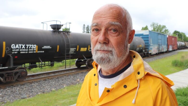 A man in a rain jacket looks at the camera. A train passes behind him carrying crude oil. 