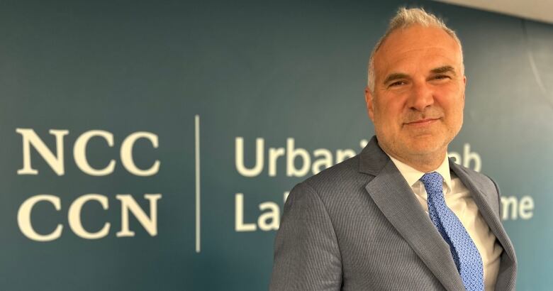 Cleanshaven man with grey hair stands with his hands in his pockets in front of a sign that reads NCC