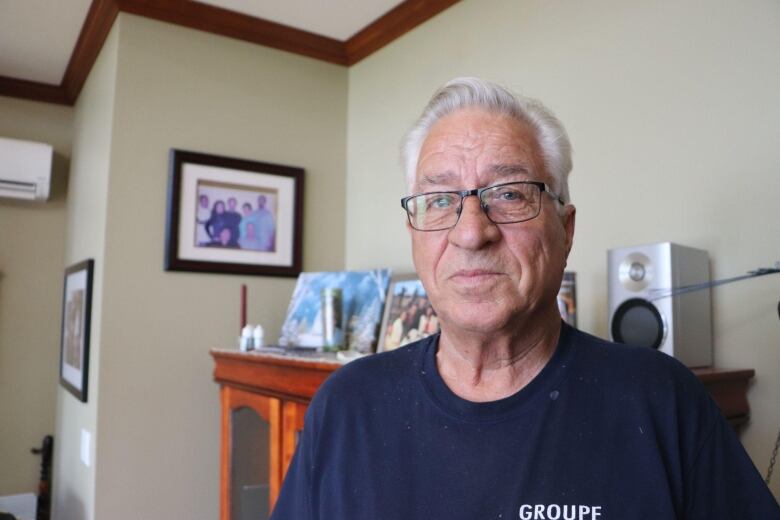 A man looks at the camera. Behind him are framed family photos. 