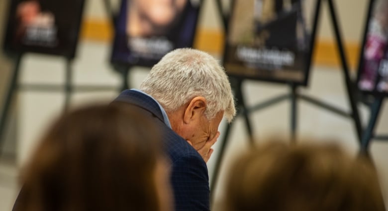 A man wearing a suit puts his head in his hands as he sits in front of a row of photos of people.