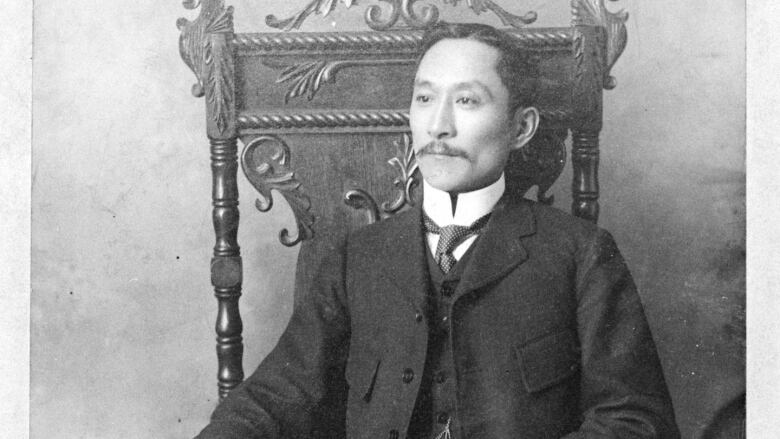 A Chinese man sits in an ornate carved chair holding a scroll. The photo is clearly old and in black and white.