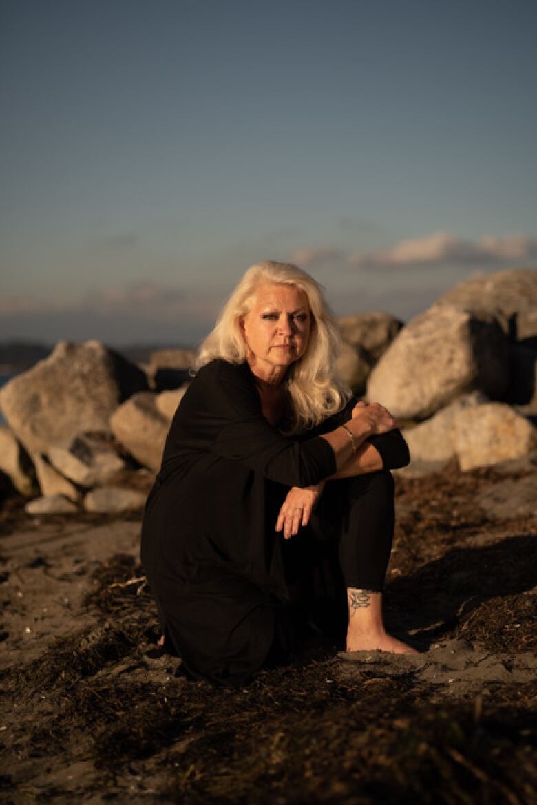 A woman sits hands on her knees on a rock with water behind.