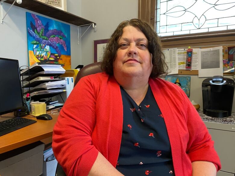 Woman in a blue shirt and red blazer sit behind a desk.