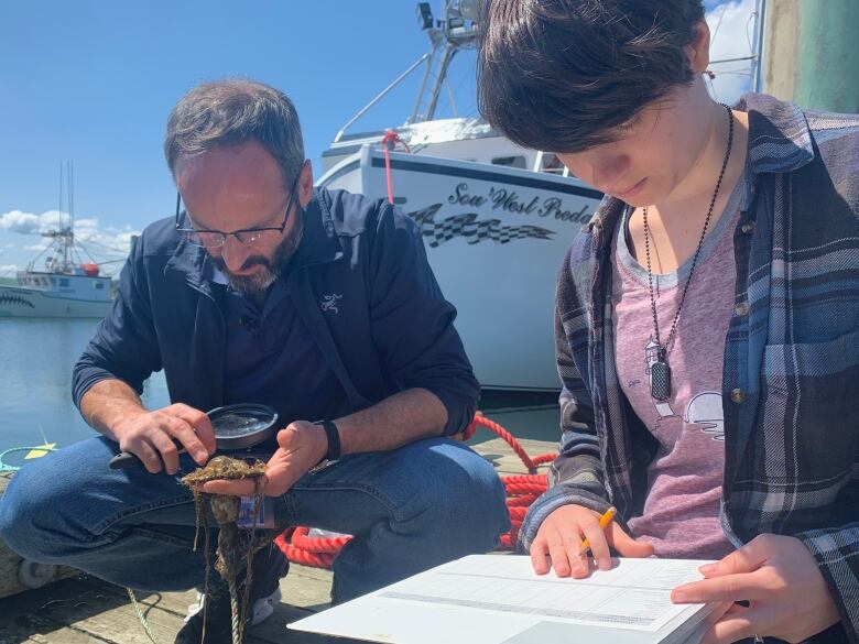 Two people set next to each other. The person on the left holds a magnifying glass over a brown, slimy blob. The person the right holds a pencil over paper.