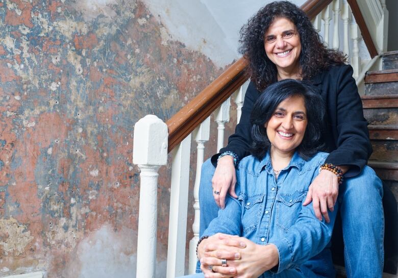 Two smiling women sit on a set of stairs, one sitting between the legs of the other.