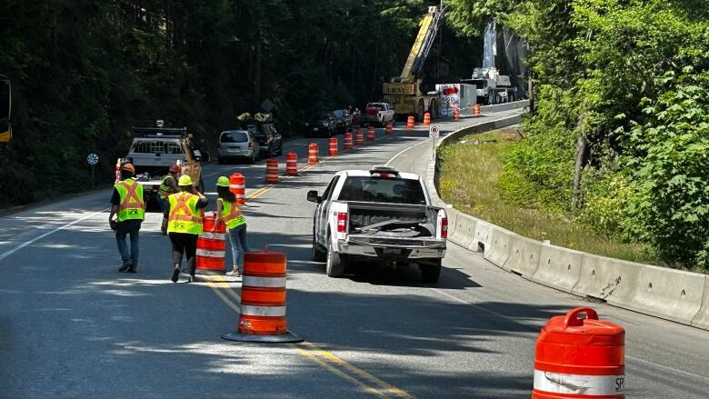 Vehicles drive on a road with officials in high visibility yellow jackets place cones between traffic lanes.