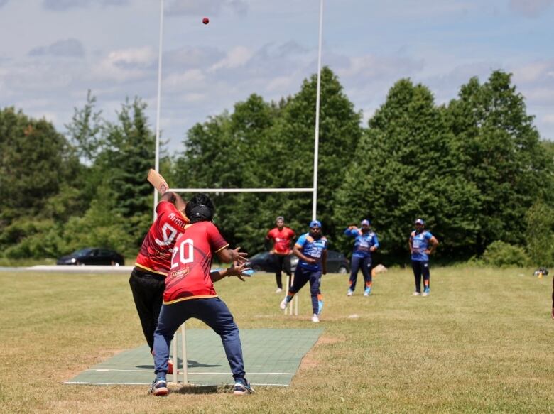 A batter standing on a critcket pitch hits the ball into the air with other players watch. 