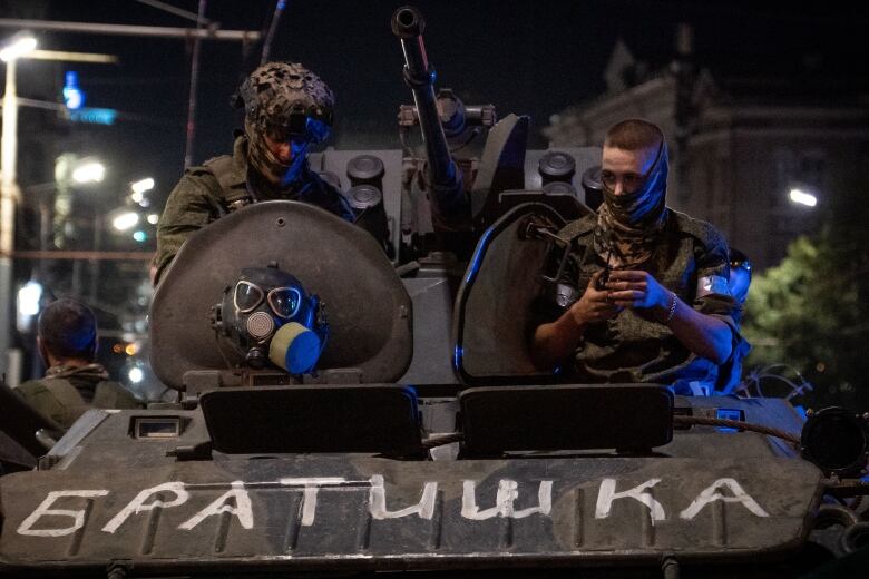 Two men in military clothing are seen on a military vehicle on an urban street.