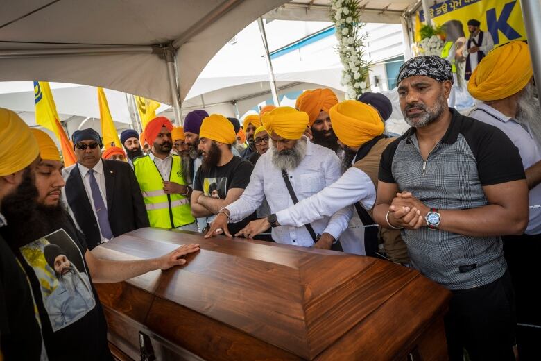 A crowd of men wearing a mix of orange and blue turbans and head coverings place their hands on a wooden casket in the centre.