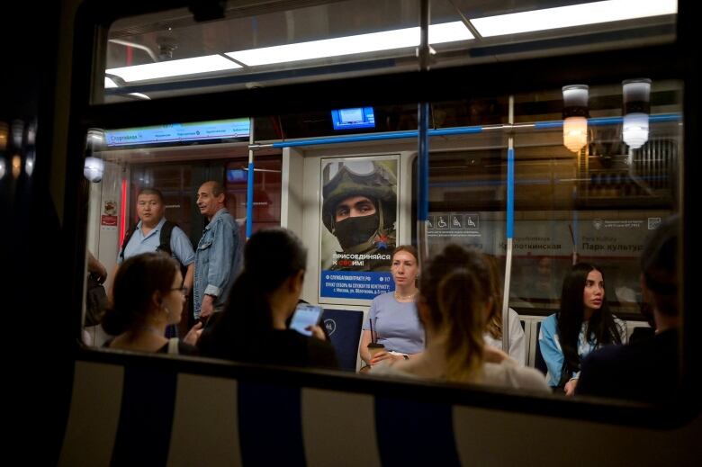 People are shown through the window of a subway, either sitting or standing. At centre is a poster of a soldier, reading 'Join your people' in Russian.