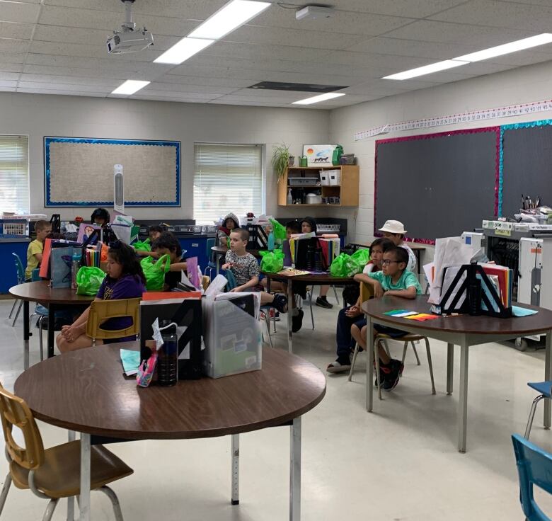 A classroom with multiple round tables has students sitting at each table with green bags of food in front of them. 