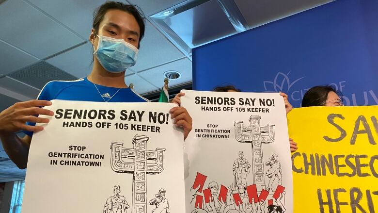 A person holds up a hoarding reading 'Seniors Say No! Hands off 105 Keefer Stop Gentrification in Chinatown' with a picture of people holding up red placards next to the Chinatown Memorial Plaza. They are next to other people holding up placards.