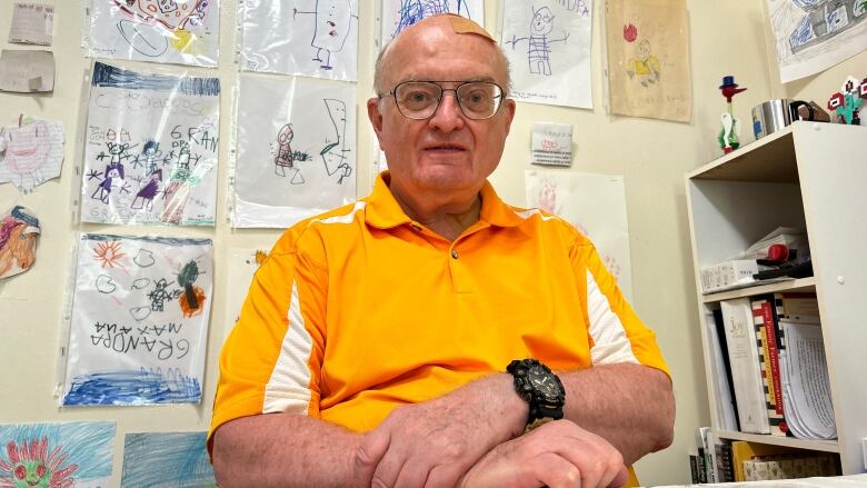 A senior wearing a bright orange shirt smiles at the camera. In the background, a wall of drawings by his grandchildren