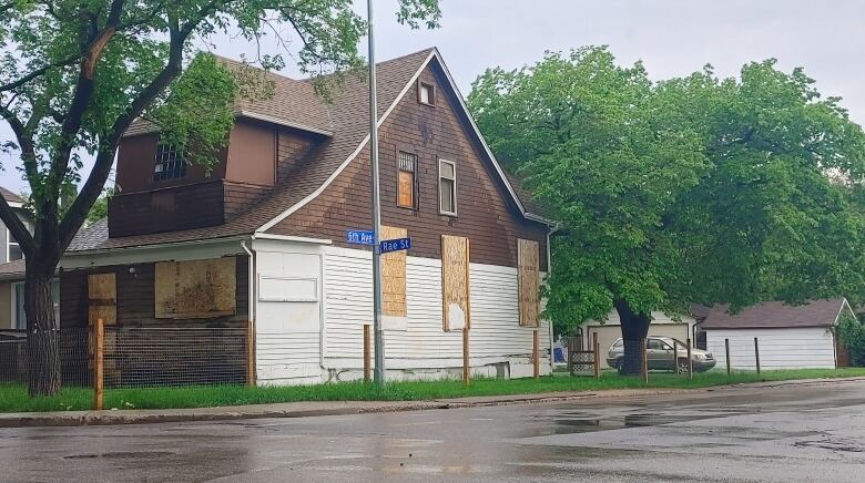 A two-storey brown-and-white house stands on the corner of a street. It is fenced in and the windows and front door are covered with plywood. A large tree is growing in the front yard of the property, and another stands in the back alley behind the house. An SUV is parked in the back alley.