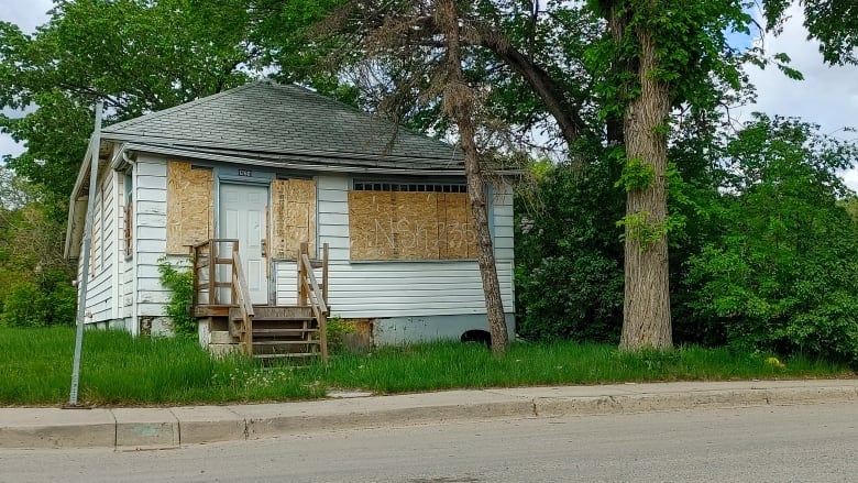 A white house with grey shingles has its windows covered by plyood. Tall green grass covers the property; tall leafy trees and shrubs hang over and beside the house.