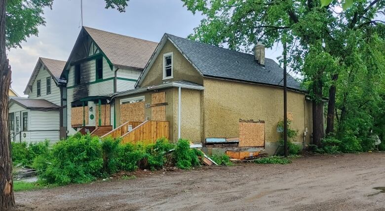 Three houses stand next to each other. One has its front windows and door placarded, as well as a hole in the right wall. The middle house is charred down the middle of its face; its windows and doors, too, are boarded up.