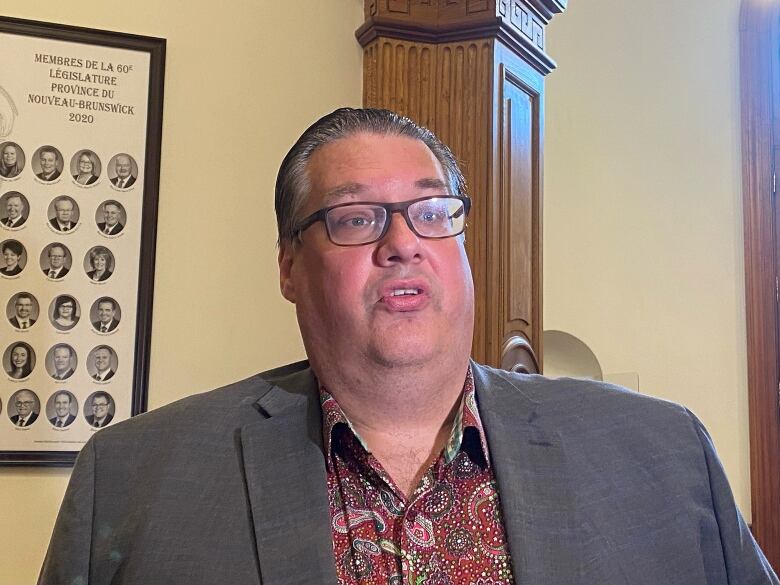 A man in glasses speaks inside a building. A photo collage of the 2020 N.B. Legislature is visible on the wall behind him.