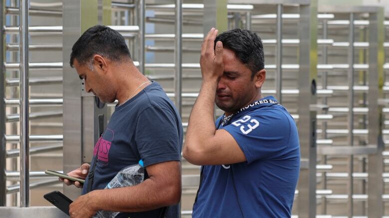 Two men wearing blue T-shirts, one in tears who is holding his hand to his face, stand outside a turnstile.