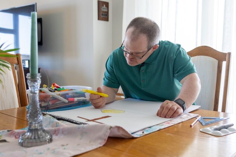 A person in a green collar shirt bends over a piece of paper on a table and works on an image of a cross.