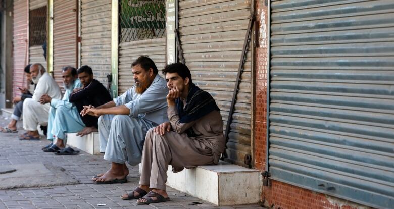 Several men sit outside closed stores.