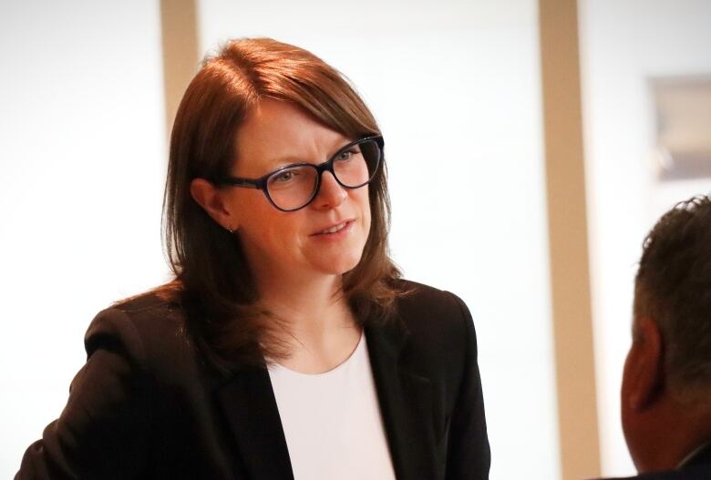 A woman with glasses and a brown sport jacket speaks with someone else.