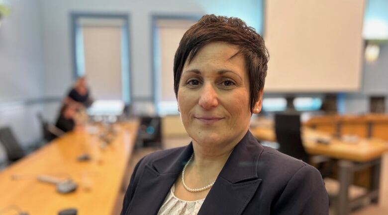 A woman in a suit looks directly at the camera as a person in the background clears up what's left over from a meeting