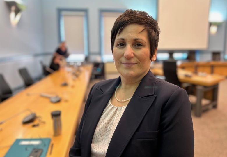 A woman in a suit looks directly at the camera as a person in the background clears up what's left over from a meeting