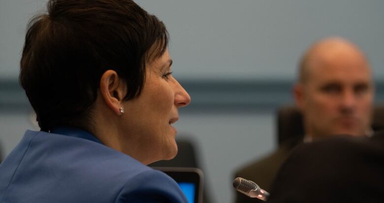 A woman with short brown hair speaks into a thin microphone at a meeting