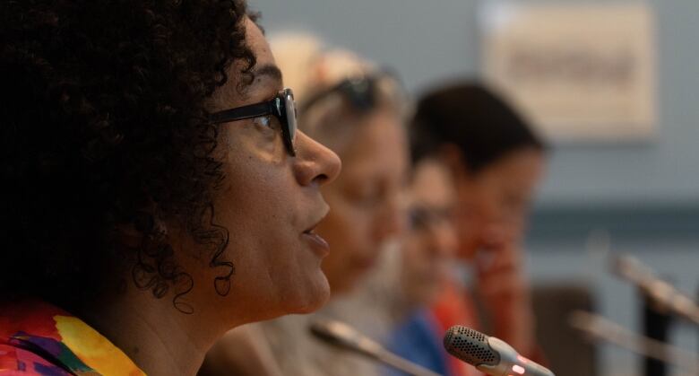 A woman with thick curly black hair and black glasses speaks into a thin microphone.