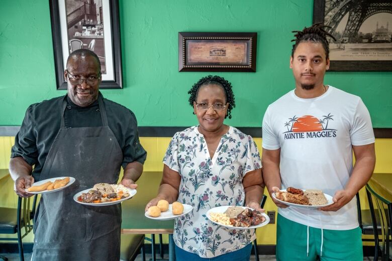 Two men, one wearing an apron and one a T-shirt reading 'Aunite Maggie's, and a woman hold plates of food in a restaurant. The wall is painted half yellow and half green like the Jamaican flag.