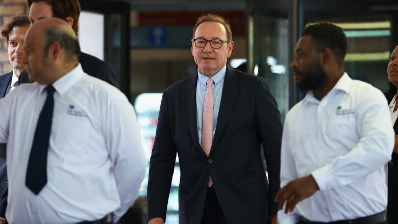 A man wears a suit, pink tie and glasses as walks outside a building in London. 