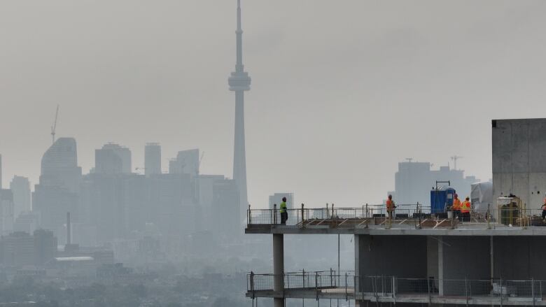 The CN Tower and Toronto skyline are hazy in the background. In the foreground construction workers stand on the upper levels of an uncompleted building.